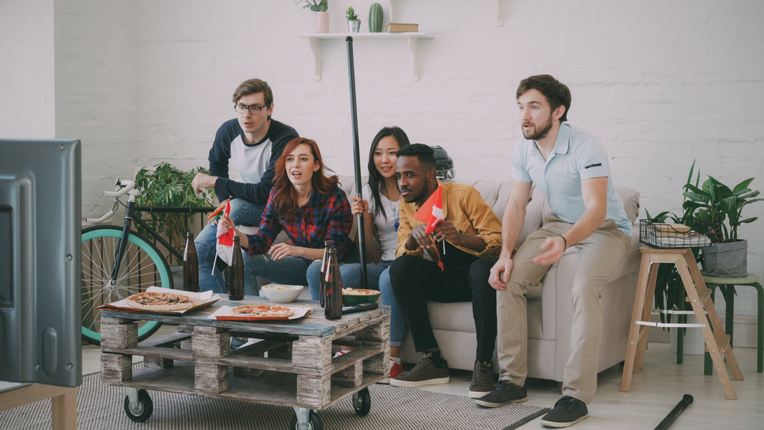 Five friends cheer on a couch while watching a game on TV, with pizza and snacks on the coffee table, capturing the game-day moment behind full-price promotions for DTC brands.