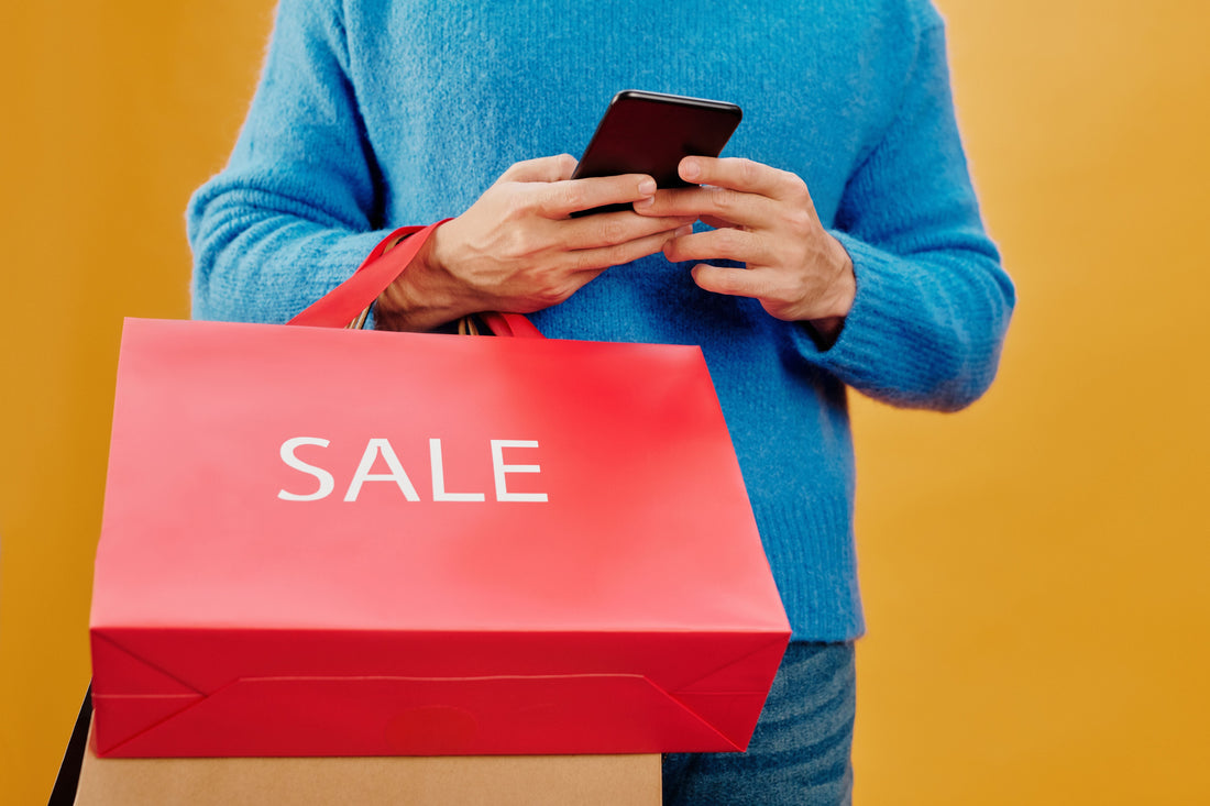 Shopper in a blue sweater texts on a phone while holding a red SALE shopping bag, illustrating the cash back vs discount code margin trade-off for Shopify brands.