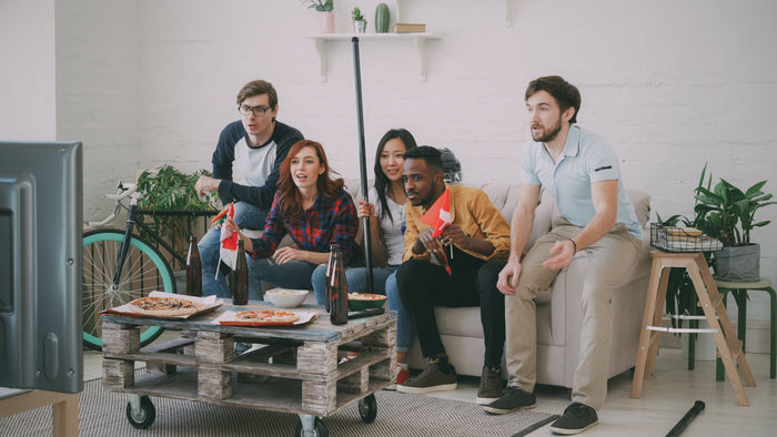 Five friends cheer on a couch while watching a game on TV, with pizza and snacks on the coffee table, capturing the game-day moment behind full-price promotions for DTC brands.