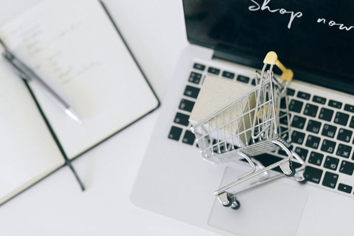 Mini shopping cart on a laptop keyboard next to a notebook, with “Shop now” on screen, for win-back campaigns