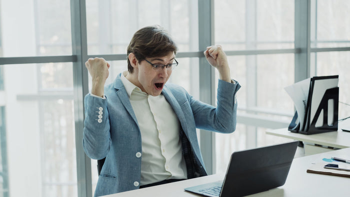 Excited man in a blazer cheering with raised fists at his laptop in a bright office, celebrating a win
